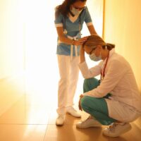 Female doctor in protective medical mask sits in corridor with her head lowered and her colleague supports her.