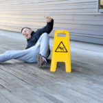 Middle-aged man yelling out in pain after falling on a slippery wooden deck right next to a bright yellow warning sign, with copy space to the side