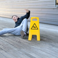 Middle-aged man yelling out in pain after falling on a slippery wooden deck right next to a bright yellow warning sign, with copy space to the side