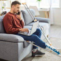 Young man with broken leg working on laptop at home talking on mobile phone sitting on sofa with crutches after accident. Male injured person using computer on rehabilitation indoors.