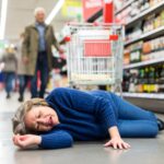 A person lies on the floor of a grocery store aisle, surrounded by shelves stocked with various beverages and a shopping cart nearby.