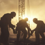 Three men in construction uniforms are working on a construction site. The sun is setting in the background, casting a warm glow over the scene. Scene is one of camaraderie and work.
