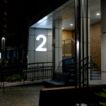 Street entrance to an apartment building with landscaping elements. A luminous number above the entrance to a modern residential complex at night.