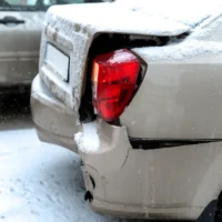 Damaged rear-end of a beige car with a broken taillight, covered in snow. Another car is partly visible in the background, conveying a winter accident scene.