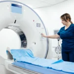 A healthcare professional in blue scrubs operates a CT scanner in a bright, sterile room. The machine's bed is covered with a blue sheet, conveying a clinical environment.