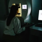 A woman in a dimly lit room sits at an eye examination machine, focused on a screen displaying an eye. A computer monitor shows test results.