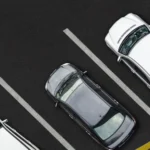 Aerial view of four parked cars in a lot, three white and one grey, neatly aligned within parking spaces. A yellow vehicle is partially visible.