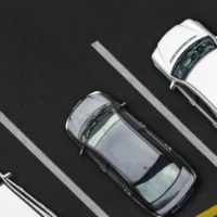 Aerial view of four parked cars in a lot, three white and one grey, neatly aligned within parking spaces. A yellow vehicle is partially visible.