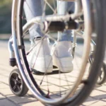 A close-up, low-angle shot looking through the large, out-of-focus wheel of a wheelchair at a person's legs and feet. The person is wearing light blue denim jeans with a frayed hem, white ribbed socks, and white canvas sneakers. They are seated on a paved brick sidewalk on a sunny day.